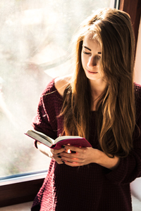 Young woman with long brown hair reading a book while standing next to a sunlit window