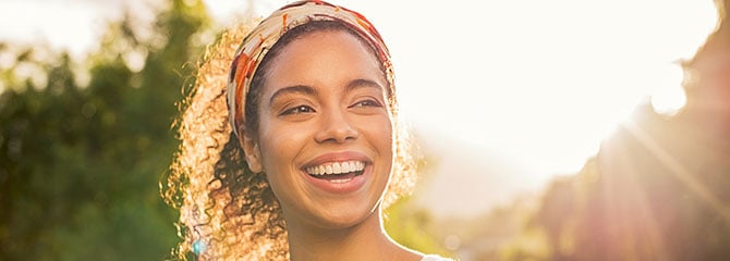 Smiling young woman wearing a tank top and headband with trees and bright sunlight behind her