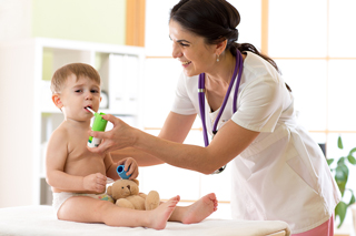 Respiratory therapist giving medicine to a toddler boy with a small teddy bear during a medical check-up in a bright clinic room