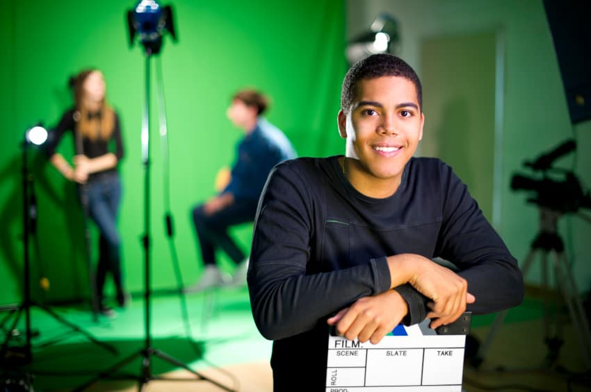 Young male production assistant smiling and holding a film slate in a studio with green screen and lighting equipment, colleagues working in the background.