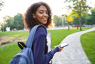 Young woman with backpack walking in park while holding smartphone