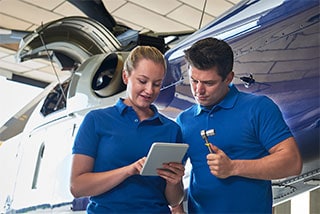 Two aviation maintenance technicians in blue shirts reviewing information on a tablet in front of an aircraft.