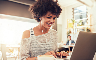 Woman with curly hair using laptop and listening to music with earphones in a bright cafe setting.