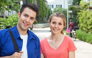 Two college students smiling outdoors, one wearing a blue shirt and backpack, the other in a pink top, with bicycles and greenery in the background.