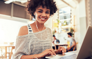 Young woman with curly hair smiling while using laptop in a cafe with earphones