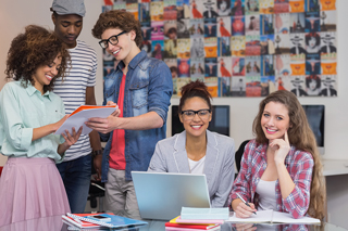 Group of five diverse young students collaborating on a project in a colorful classroom with laptops and notebooks, with a poster-filled wall in the background.