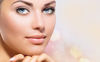Close-up of a woman with clear skin and striking blue eyes, resting her chin on her hands with a soft focus background.