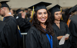Female graduate smiling at graduation ceremony, wearing black cap and gown with a blue and gold honor cord.