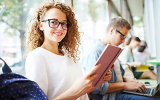 Curly-haired woman with glasses reading a book in a bustling cafe with other people working in the background