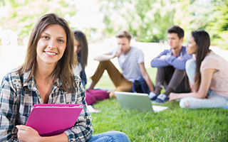 Young woman holding a notebook with a group of students sitting in the grass in the background at a park.