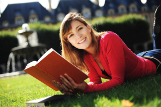 Young woman reading a book on grass in sunny outdoor setting with buildings in background