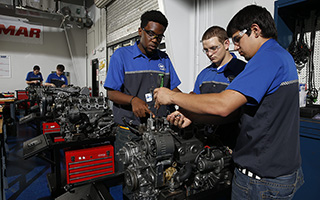 Three young male engineers working on machinery in an automotive workshop, wearing blue uniforms.