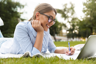 Smiling young woman with glasses lying on a blanket in a grassy field while holding a pen and looking at a laptop screen