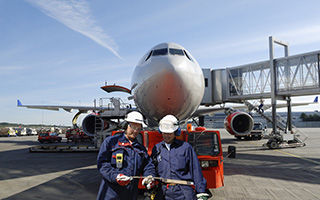 Two airport ground staff in blue uniforms and helmets discussing in front of a parked airplane at an airport terminal with jet bridge and equipment around.