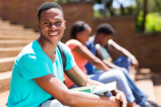 Happy young man holding a book sitting on outdoor stairs at university campus with other students in the background