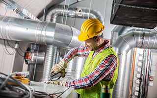 Male worker in a reflective vest and yellow helmet inspecting or repairing industrial HVAC system pipes in a mechanical room.