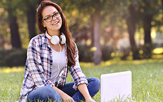 Young woman with glasses sitting on grass in park with laptop and headphones, smiling at camera.