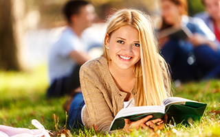 Young woman reading a book outdoors at a park with other students in the background