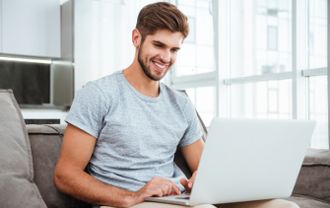 Smiling young man using laptop while sitting on couch in modern living room.