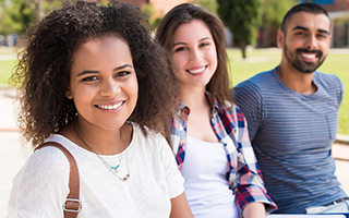 Three cheerful young adults sitting outdoors on a sunny day, with a woman in the forefront smiling at the camera and two others in the background, also smiling.