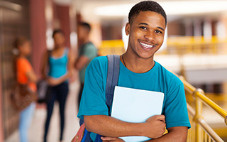 Smiling teenage boy with backpack holding a notebook in school hallway