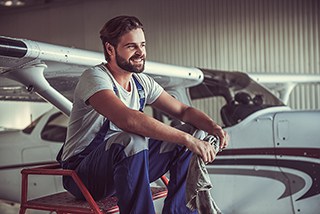 Man with beard sitting on a ladder next to a small airplane in a hangar