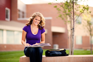 Young woman studying outdoors on a college campus, sitting on a bench with books and a backpack, wearing a purple shirt