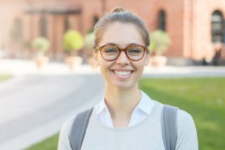 Happy young woman with glasses smiling at the camera in front of a brick building on a sunny day.