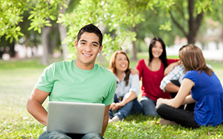 Young man with laptop sitting on grass in park with three friends in background smiling and enjoying sunny day