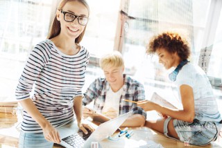 Smiling woman with glasses using laptop with two colleagues discussing paperwork in a bright office setting