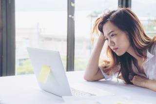 Woman looking tired while working on laptop in a bright office setting.