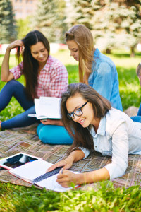 Three female students studying together outdoors on a blanket with books and a laptop