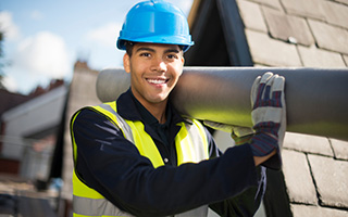 Construction worker with blue hard hat and high visibility jacket carrying a large metal pipe outdoors