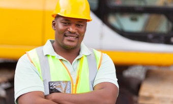 Smiling man wearing a yellow hard hat and bright safety vest standing outside in front of heavy construction equipment