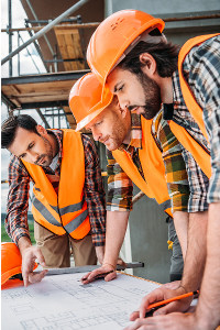 Three construction workers in orange hard hats and reflective vests analyzing blueprints at a construction site.