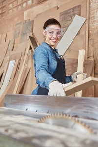 Woman carpenter in safety goggles and gloves cutting wood with a table saw in a workshop.