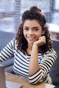 Young woman with curly hair wearing a striped top sitting at a desk with a laptop in an office setting