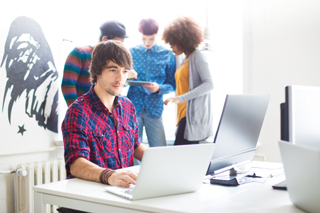 Software engineer in a plaid shirt working on a laptop in a bright office with colleagues discussing in the background.