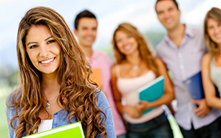 Young woman with long brown hair holding books, standing in foreground with a group of smiling friends in casual clothing in the background.