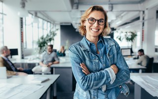 Woman with glasses and denim jacket standing confidently in busy office environment