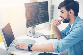Young male programmer coding on laptop and desktop with code on screens in a bright office.