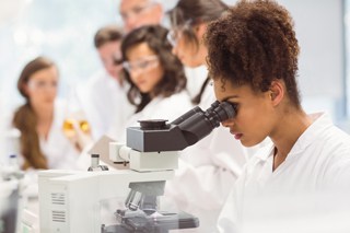 Young female scientist using microscope in laboratory with colleagues working in background