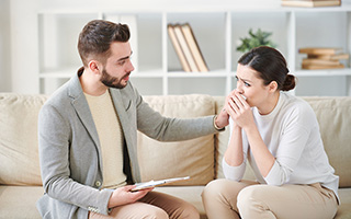 Man comforting upset woman on sofa while holding a document in a counseling office.