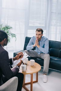 Two men in a serious discussion in an office, one sitting with hands clasped reflecting and the other holding documents, with coffee and a smartphone on a small table between them.