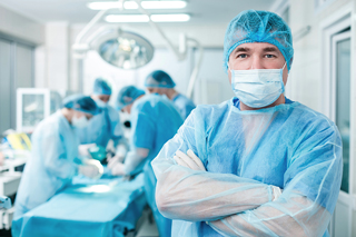 Surgical technician n in blue scrubs and surgical mask standing with arms crossed in operating room with medical team performing surgery in background.