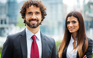 Two professional business people, a man with curly hair smiling in a suit and red tie, and a woman with long brown hair in a gray suit, standing outside office buildings.