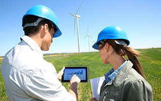 Two engineers with helmets analyzing data on a tablet in front of wind turbines in a field on a sunny day.