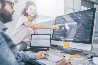 Two software developers, a man with a beard and glasses and a woman with curly hair, collaborating at a workstation with multiple monitors displaying code and data analysis.