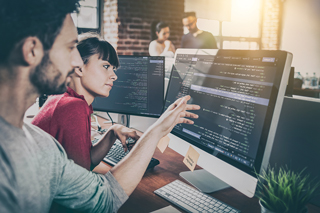 Two software developers collaborating at a workstation with dual monitors displaying code, in an office setting with colleagues in the background.