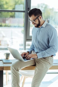 Man with glasses sitting partially on a wooden desk in front of large windows and typing on an open laptop on his leg